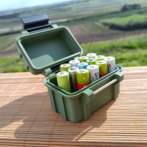 Könnte beinhalten: Eine grüne Aufbewahrungsbox aus Kunststoff mit einem schwarzen Verschluss, gefüllt mit verschiedenen AA-Batterien. Die Box hat einen Metallgriff und steht auf einer geflochtenen Bambusmatte.