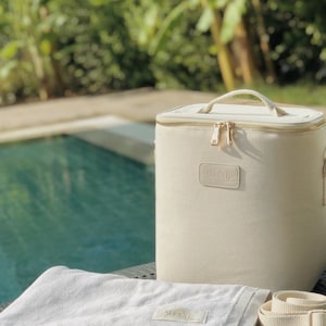 May include: A white canvas cooler bag with a gold zipper and a gold handle. The bag has the word "SUMMER" printed on it in gold lettering. The bag is sitting on a wooden table next to a white towel with a fringe border and a white canvas strap.