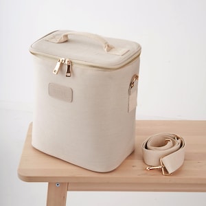 May include: A beige canvas cooler bag with a gold zipper and a gold handle. The bag has a gold metal buckle and a removable beige canvas strap. The bag is sitting on a wooden table.
