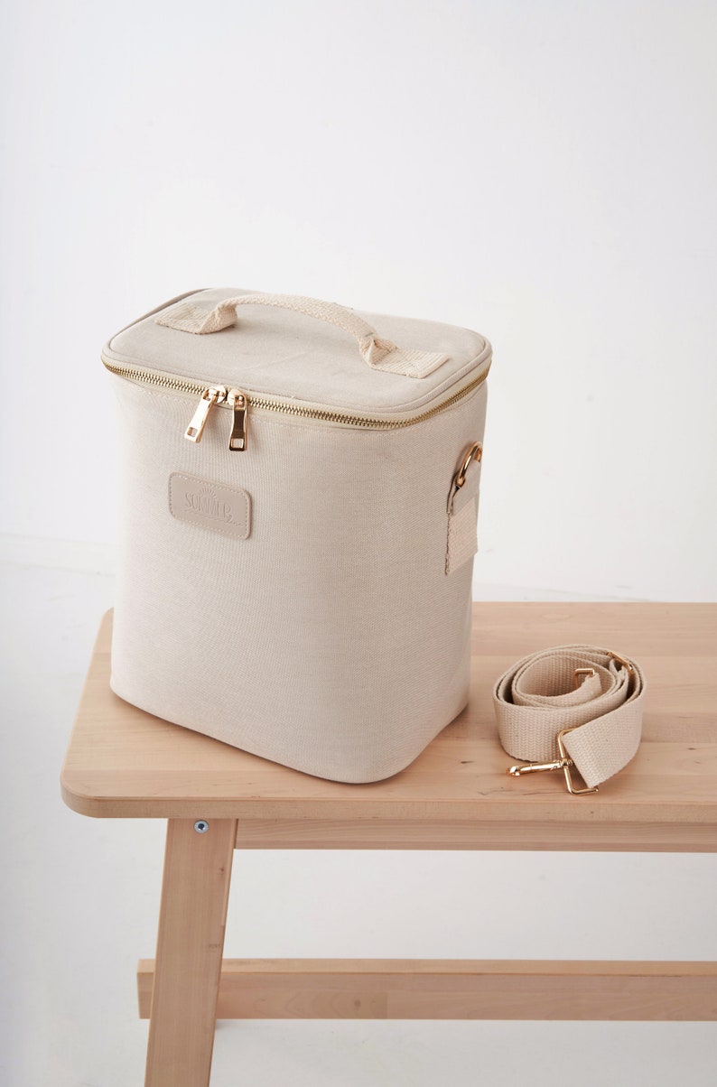 May include: A beige canvas cooler bag with a gold zipper and a gold handle. The bag has a gold metal buckle and a removable beige canvas strap. The bag is sitting on a wooden table.