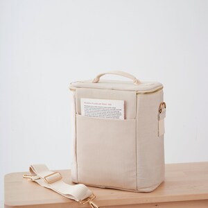 May include: A beige canvas lunch bag with a gold zipper and a gold handle. The bag has a small pocket on the front with a piece of paper inside that says "MODERN KLASSIKER DUFT - 60". The bag has a long beige canvas strap with gold hardware.
