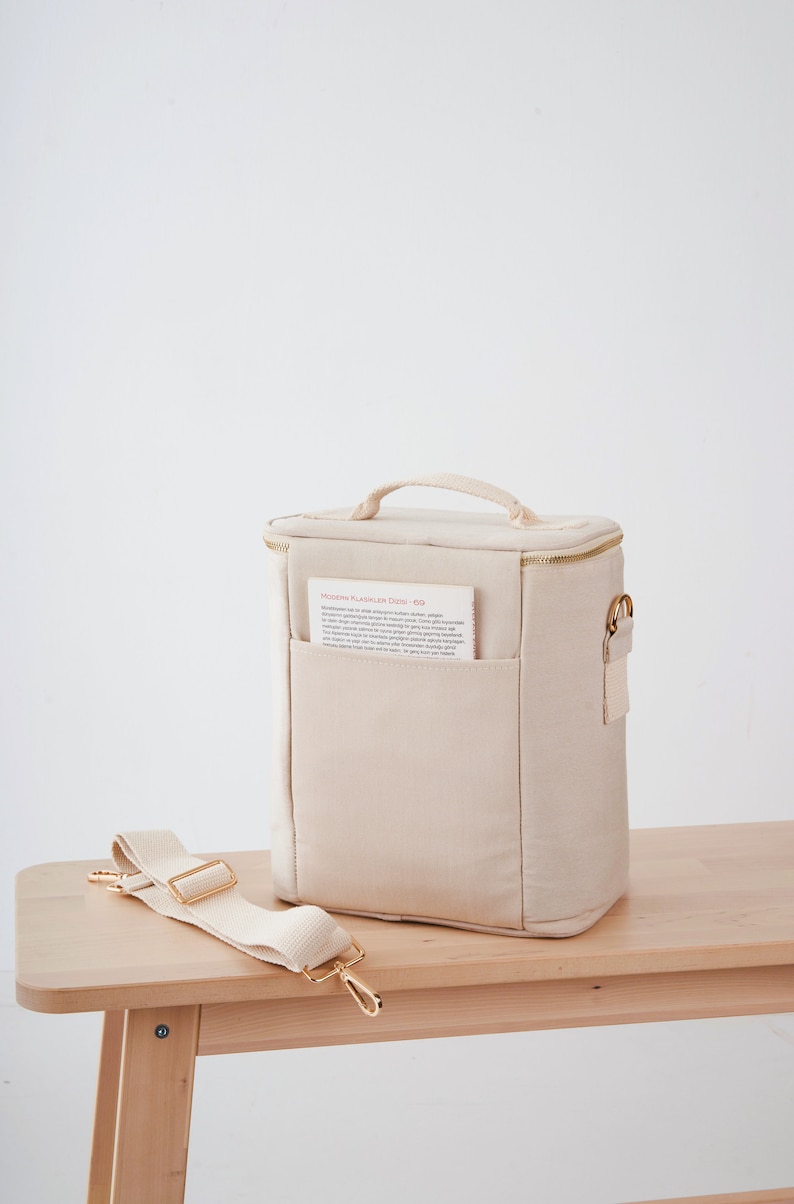 May include: A beige canvas lunch bag with a gold zipper and a gold handle. The bag has a small pocket on the front with a piece of paper inside that says "MODERN KLASSIKER DUFT - 60". The bag has a long beige canvas strap with gold hardware.