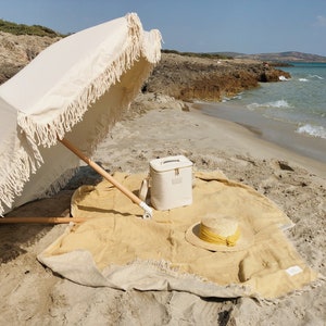May include: A white beach umbrella with fringe trim, a white cooler bag, a straw hat with a yellow ribbon, and a yellow and beige beach blanket on a sandy beach.