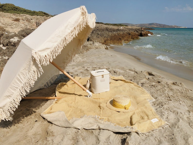 May include: A white beach umbrella with fringe trim, a white cooler bag, a straw hat with a yellow ribbon, and a yellow and beige beach blanket on a sandy beach.