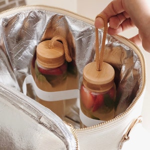 May include: A white insulated lunch bag with a zipper closure and a gold handle. The bag contains two glass bottles with wooden lids and a straw. The bottles are filled with a clear liquid and red fruit.