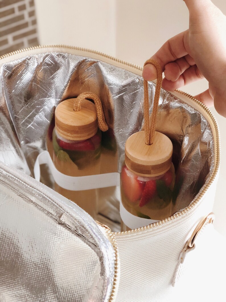 May include: A white insulated lunch bag with a zipper closure and a gold handle. The bag contains two glass bottles with wooden lids and a straw. The bottles are filled with a clear liquid and red fruit.