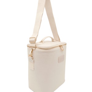 May include: A white canvas lunch bag with a gold zipper and a gold strap. The bag has a rectangular shape and a small patch on the front.