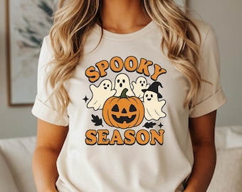May include: Beige t-shirt with the words "Spooky Season" in orange, featuring a jack-o'-lantern, ghosts, and a witch's hat. The design is in a retro style, perfect for Halloween.