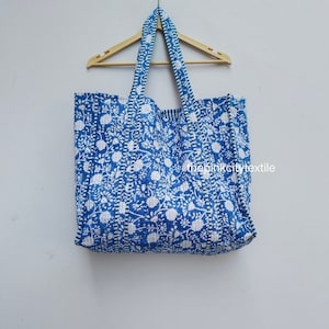 Op de afbeelding: Een grote blauwe tote bag met een witte bloemenprint en witte bies. De tas heeft twee lange hengsels en hangt aan een houten hanger.