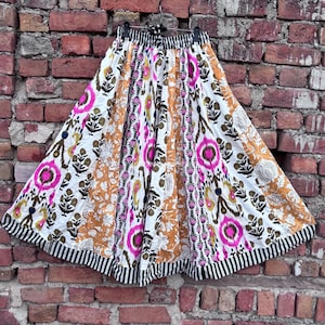 Könnte beinhalten: Ein langer, fließender Rock mit einem lebendigen, böhmischen Design. Der Rock hat eine weiße Basis mit einem sich wiederholenden Muster aus rosa, braunen und orangefarbenen Blumen- und geometrischen Formen. Der Saum ist mit einem schwarz-weiß gestreiften Rand versehen.