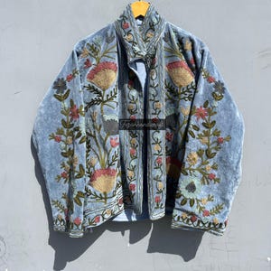 Può includere: Una giacca di velluto blu chiaro con ricami floreali. I ricami sono in tonalità di rosa, verde e giallo. La giacca ha un colletto alla coreana e una chiusura con bottoni.