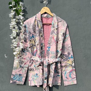 May include: A pink quilted jacket with a floral print and a tie belt. The jacket has long sleeves and a V-neck. The print features birds, leaves, and other floral elements.