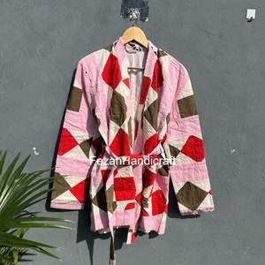 Peut inclure: Veste kimono rose, rouge et marron à motifs géométriques avec une ceinture assortie. La veste est suspendue à un cintre en bois contre un mur gris. Le motif de la veste présente un patchwork de losanges et de carrés.