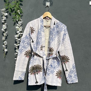 Puede incluir: Chaqueta de algodón blanca con estampado floral azul y marrón. La chaqueta tiene un cinturón de lazo y mangas largas.