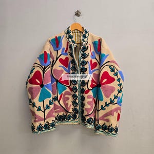 以下が含まれることがあります： カラフルな花の刺繍が施されたベージュのジャケット。 赤、青、ピンクの花と緑の葉と茎が特徴です。 ジャケットは、ニュートラルな背景に木製のハンガーに吊るされています。