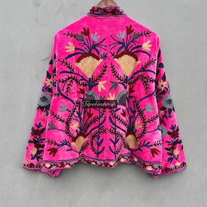 Puede incluir: Una chaqueta de terciopelo rosa con intrincados bordados florales en tonos azules, rojos y dorados. La chaqueta tiene un cuello alto y mangas largas. "Tezjanhandicraft" est&aacute; bordado en la espalda.