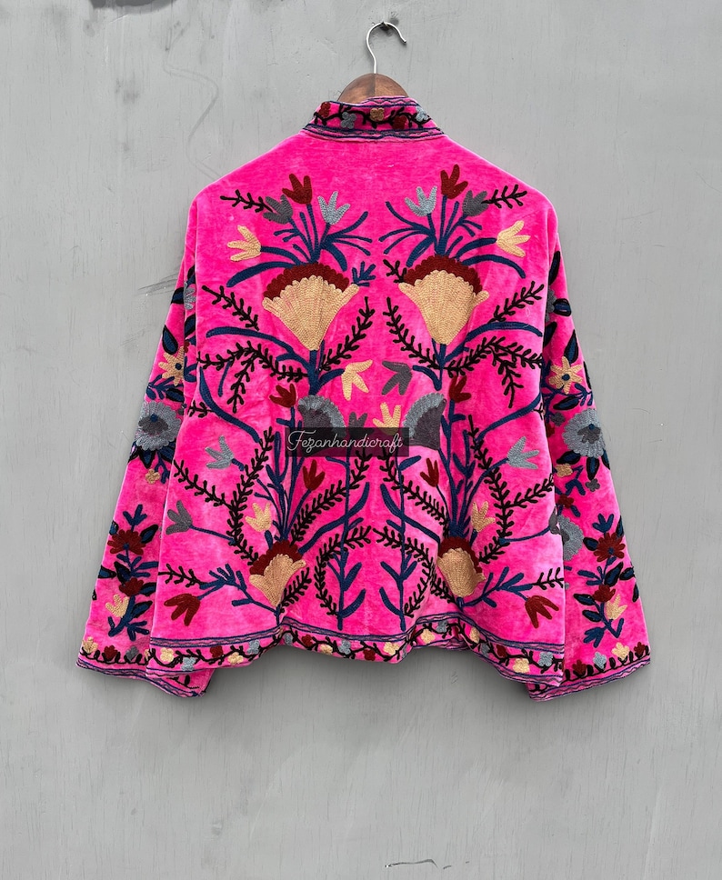 Puede incluir: Una chaqueta de terciopelo rosa con intrincados bordados florales en tonos azules, rojos y dorados. La chaqueta tiene un cuello alto y mangas largas. "Tezjanhandicraft" est&aacute; bordado en la espalda.