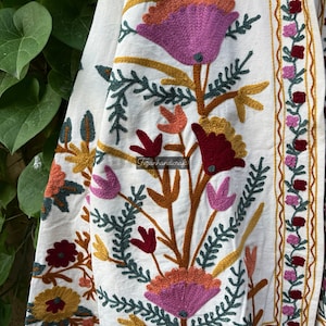 May include: White cotton fabric with embroidered floral designs in shades of pink, red, orange, and green. The fabric has a border with a wavy pattern.