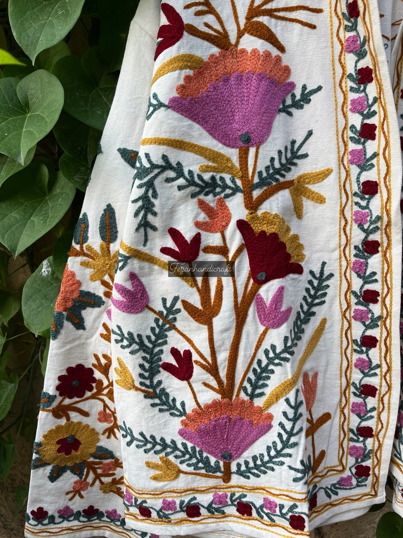 May include: White cotton fabric with embroidered floral designs in shades of pink, red, orange, and green. The fabric has a border with a wavy pattern.