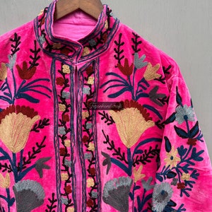 Puede incluir: Una chaqueta de terciopelo rosa con intrincados bordados florales en tonos de azul, rojo, dorado y gris. La chaqueta tiene un cuello mandar&iacute;n y un cierre frontal.
