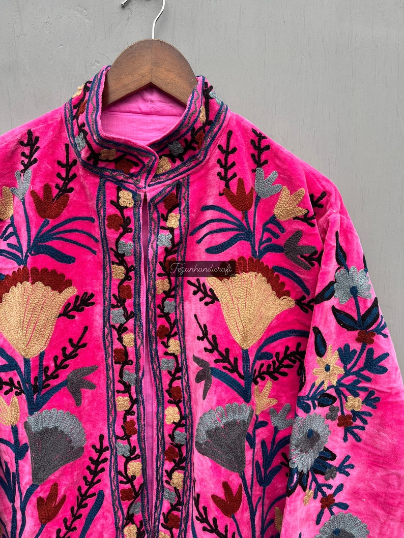 Puede incluir: Una chaqueta de terciopelo rosa con intrincados bordados florales en tonos de azul, rojo, dorado y gris. La chaqueta tiene un cuello mandar&iacute;n y un cierre frontal.