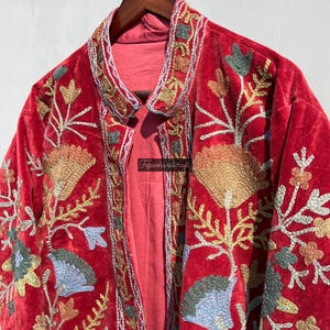 May include: A red velvet jacket with intricate floral embroidery in gold, blue, and green thread. The jacket has a mandarin collar and a long, flowing silhouette.