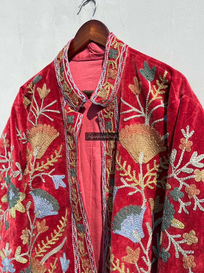 May include: A red velvet jacket with intricate floral embroidery in gold, blue, and green thread. The jacket has a mandarin collar and a long, flowing silhouette.