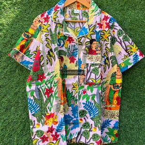 Puede incluir: Una camisa rosa de botones con estampado floral colorido con im&aacute;genes de Frida Kahlo. La camisa tiene un cuello blanco y un ribete blanco alrededor de las mangas y el cuello.