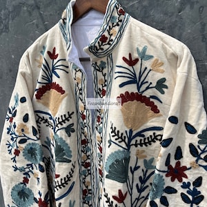 May include: A cream-colored jacket with intricate floral embroidery in shades of blue, red, and gold. The embroidery features a variety of floral motifs, including leaves, flowers, and stems.
