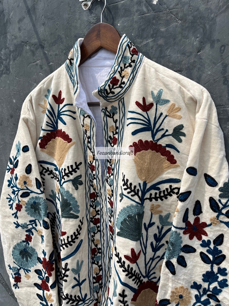 May include: A cream-colored jacket with intricate floral embroidery in shades of blue, red, and gold. The embroidery features a variety of floral motifs, including leaves, flowers, and stems.