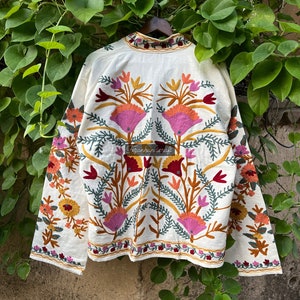 May include: A white cotton jacket with colorful floral embroidery. The embroidery features pink, orange, yellow, green, and red thread. The jacket has long sleeves and a high neck.