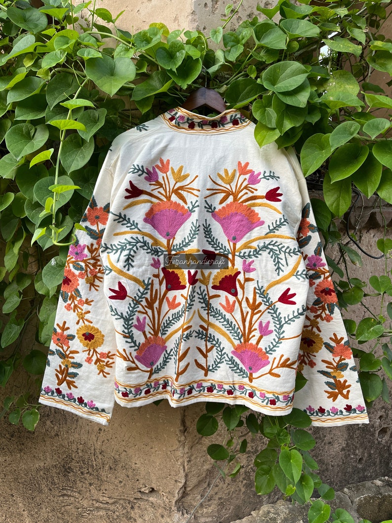 May include: A white cotton jacket with colorful floral embroidery. The embroidery features pink, orange, yellow, green, and red thread. The jacket has long sleeves and a high neck.
