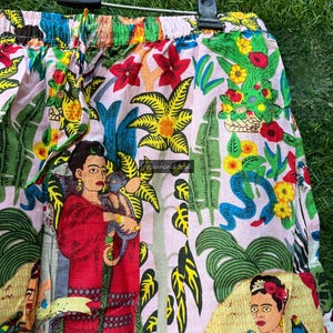 Puede incluir: Shorts de algod&oacute;n rosa con un estampado floral colorido que presenta una imagen repetida de Frida Kahlo. El estampado tambi&eacute;n incluye plantas tropicales, serpientes y loros.