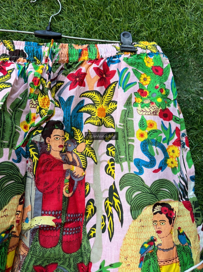 Puede incluir: Shorts de algod&oacute;n rosa con un estampado floral colorido que presenta una imagen repetida de Frida Kahlo. El estampado tambi&eacute;n incluye plantas tropicales, serpientes y loros.