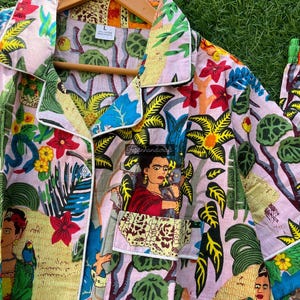 Puede incluir: Una camisa de botones rosa con ribete blanco. La camisa presenta un estampado floral colorido con im&aacute;genes de Frida Kahlo y loros.