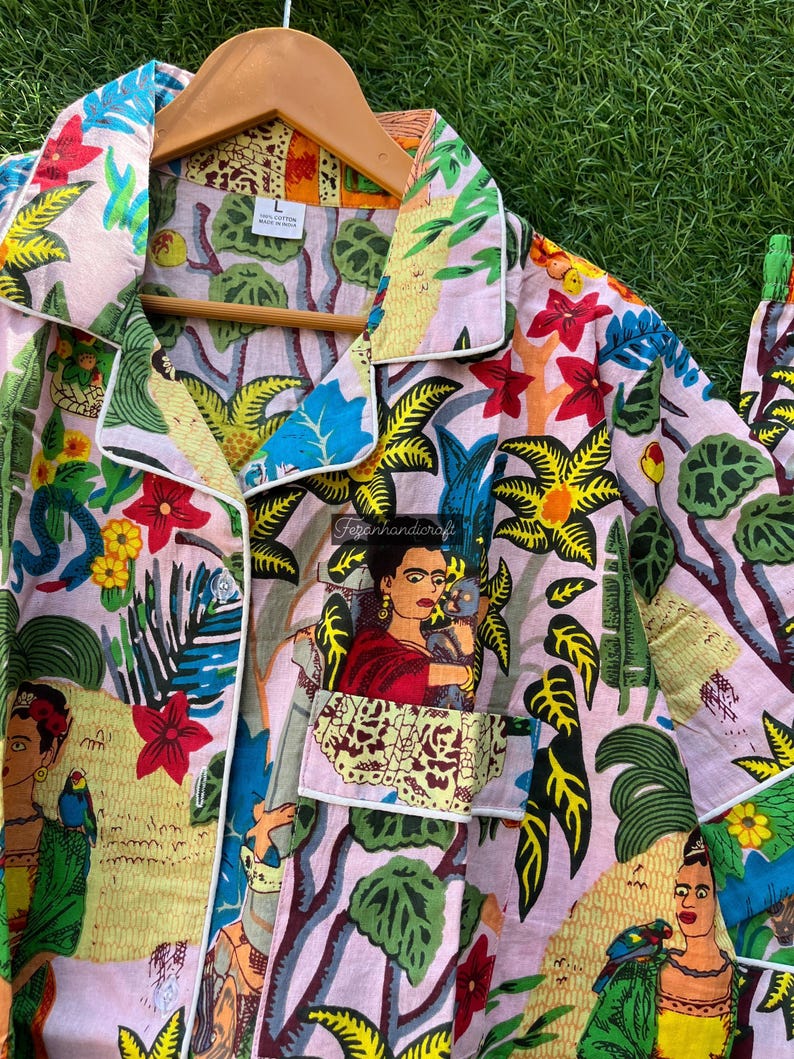 Puede incluir: Una camisa de botones rosa con ribete blanco. La camisa presenta un estampado floral colorido con im&aacute;genes de Frida Kahlo y loros.