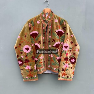 May include: A mustard velvet jacket with detailed floral embroidery in pink, burgundy, and orange. The jacket has a mandarin collar and is hung on a wooden hanger. The text "Fezanhandicraft" is visible on the jacket.