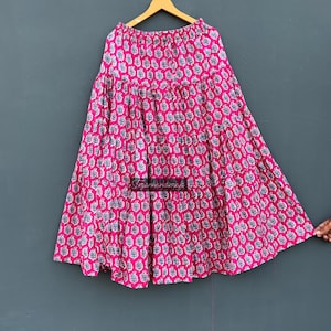 Puede incluir: Una falda rosa vibrante con un diseño escalonado, adornada con un patrón repetido de motivos florales blancos y azules. La falda está colgada en una percha de madera sobre un fondo gris. La tela de la falda parece ligera.