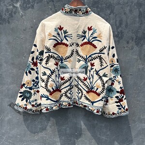 May include: A white cotton jacket with colorful floral embroidery. The embroidery features a symmetrical design with red, blue, and yellow flowers.