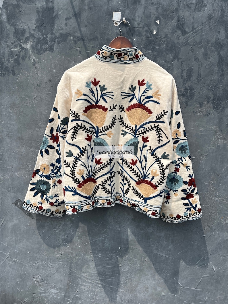 May include: A white cotton jacket with colorful floral embroidery. The embroidery features a symmetrical design with red, blue, and yellow flowers.