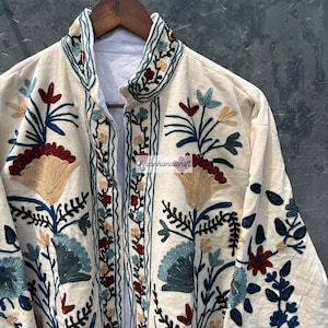 May include: A white cotton jacket with intricate floral embroidery in shades of blue, red, and gold. The jacket has a mandarin collar and a front closure.