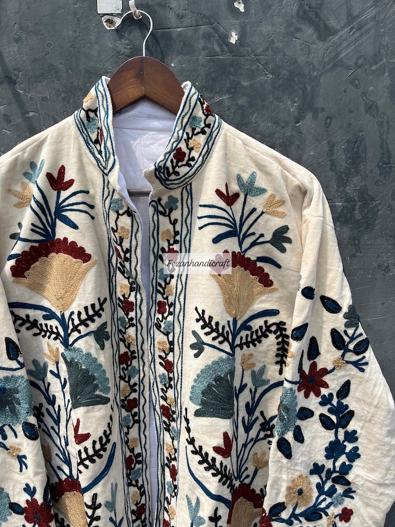 May include: A white cotton jacket with intricate floral embroidery in shades of blue, red, and gold. The jacket has a mandarin collar and a front closure.