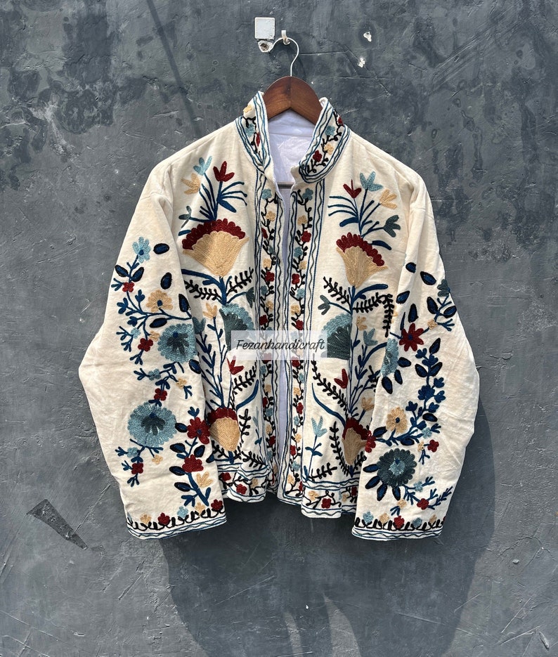 May include: A white cotton jacket with a mandarin collar and long sleeves. The jacket is embroidered with colorful floral patterns in shades of blue, red, yellow, and green. The embroidery is intricate and detailed, and the jacket has a vintage look.