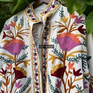 May include: A white cotton jacket with colorful floral embroidery. The embroidery features pink, orange, yellow, green, and burgundy flowers. The jacket has a mandarin collar and a button closure.