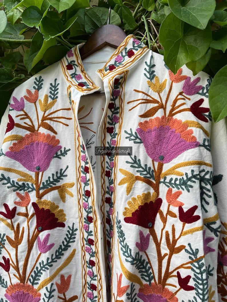 May include: A white cotton jacket with colorful floral embroidery. The embroidery features pink, orange, yellow, green, and burgundy flowers. The jacket has a mandarin collar and a button closure.