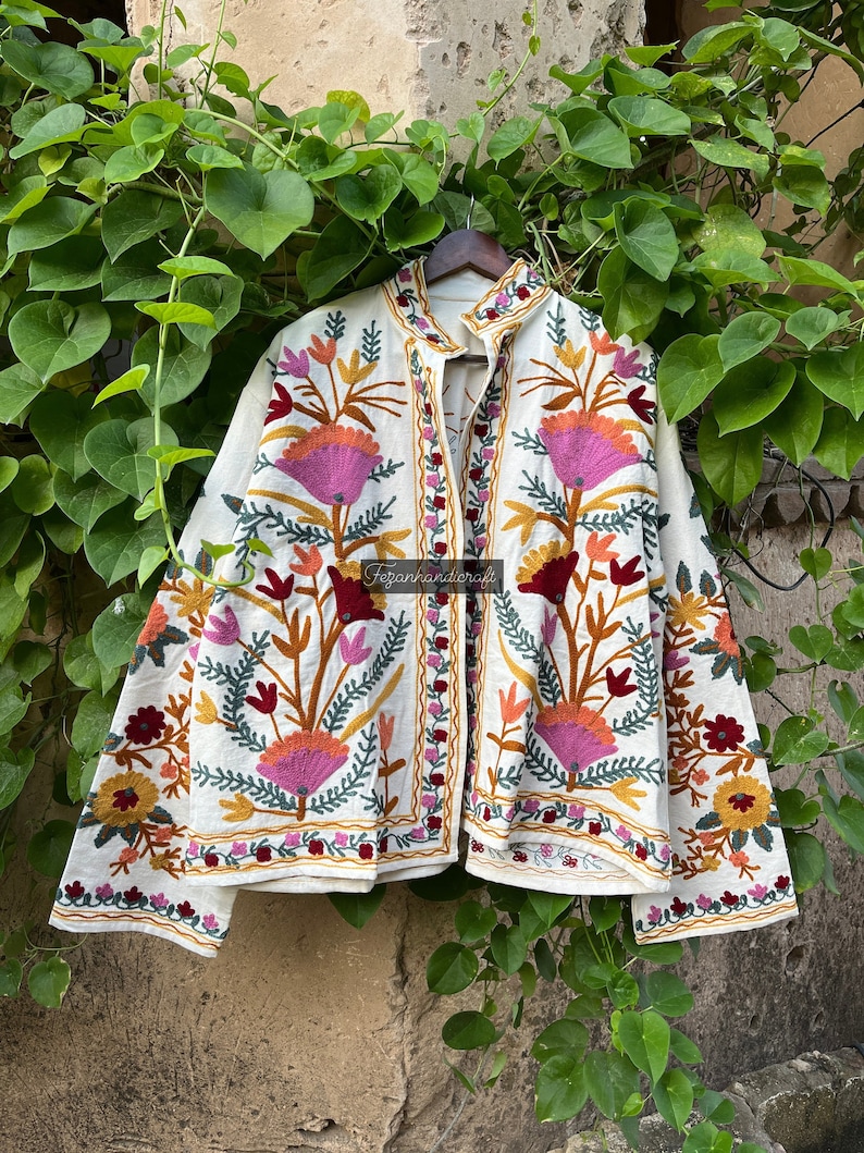 May include: A white embroidered jacket with floral patterns in shades of pink, orange, yellow, green, and red. The jacket has a mandarin collar and a button closure.
