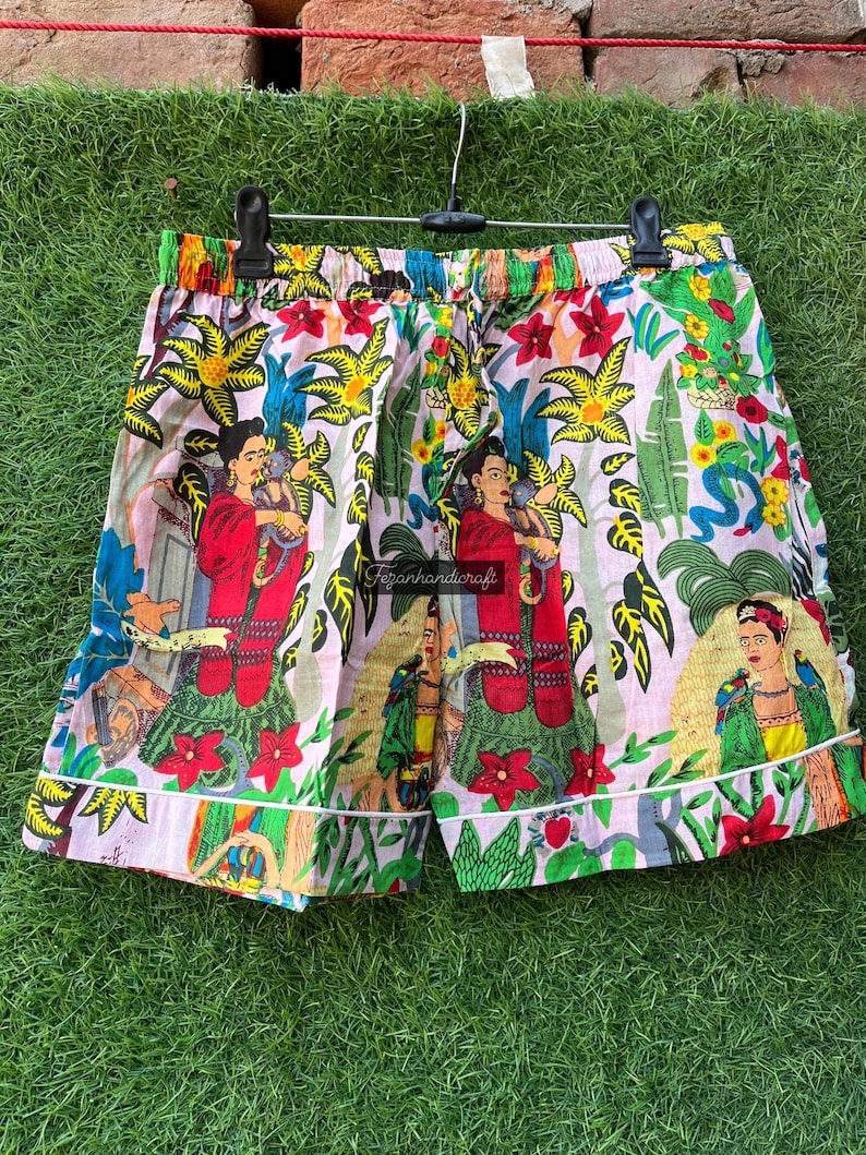 Puede incluir: Un par de pantalones cortos de algod&oacute;n rosa y blanco con un estampado floral colorido que presenta una imagen repetida de una mujer con una flor en el pelo. El estampado tambi&eacute;n incluye loros, flores y hojas.