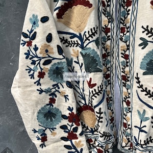 May include: A white cotton jacket with intricate floral embroidery in shades of blue, green, red, and beige. The embroidery features a variety of flowers and leaves, creating a beautiful and intricate design.