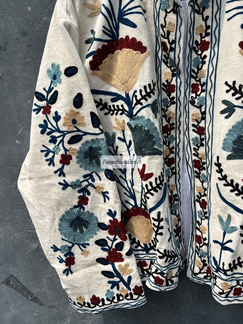 May include: A white cotton jacket with intricate floral embroidery in shades of blue, green, red, and beige. The embroidery features a variety of flowers and leaves, creating a beautiful and intricate design.
