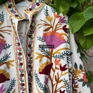 May include: A white cotton jacket with colorful floral embroidery. The embroidery features pink, red, orange, and yellow flowers with green leaves. The jacket has a mandarin collar and a front closure.
